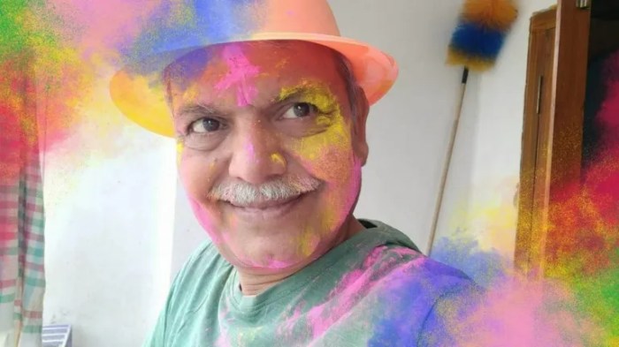 A smiling man wearing a yellow hat and a green shirt, with colorful powder smeared on his face, celebrating a festival.