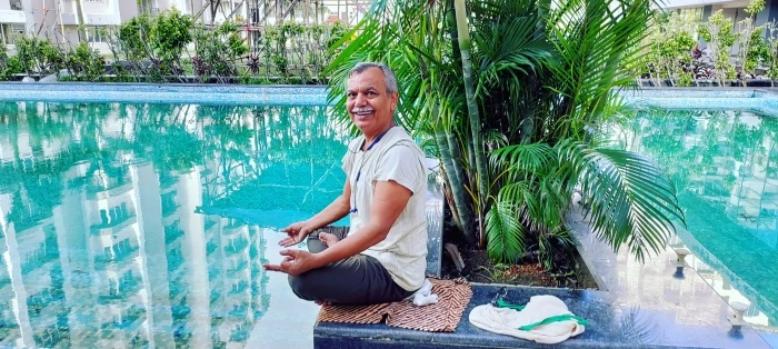 A smiling man sitting near a tranquil swimming pool, surrounded by greenery, practicing meditation.