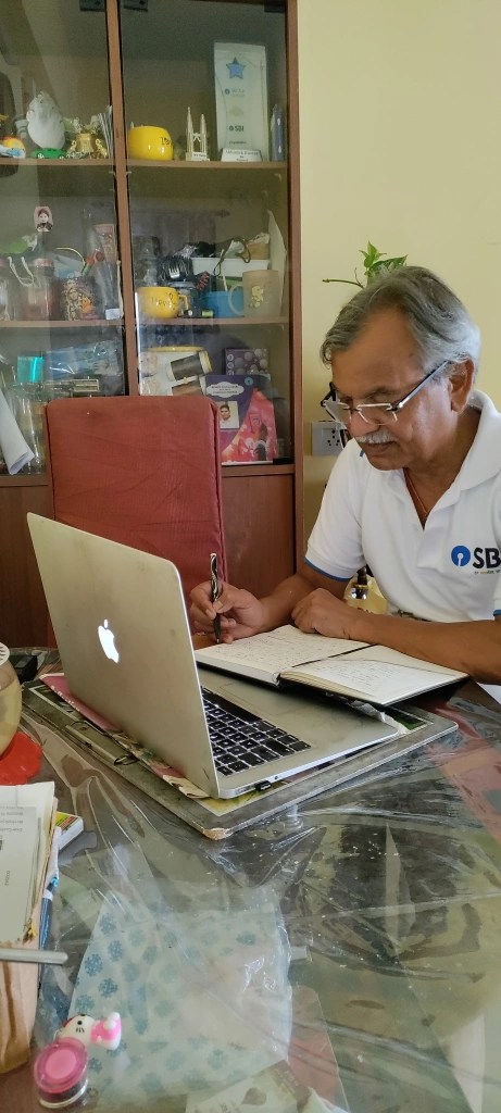 A man sitting at a table with a laptop and a notebook, writing notes. A display cabinet with various items is in the background.