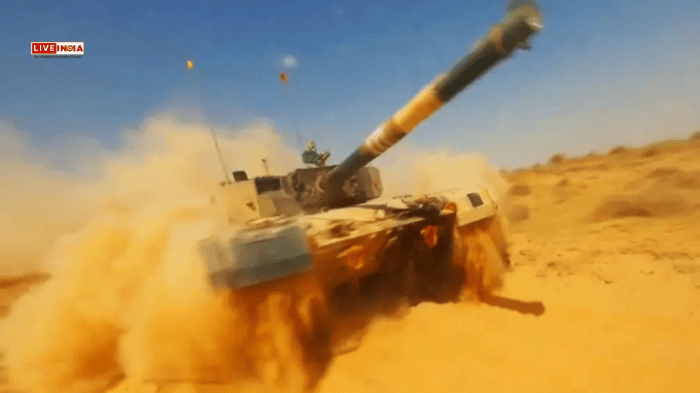 A military tank is driving through a dusty terrain, creating a cloud of dust. The tank's turret is raised, and a soldier can be seen atop it against a clear blue sky.